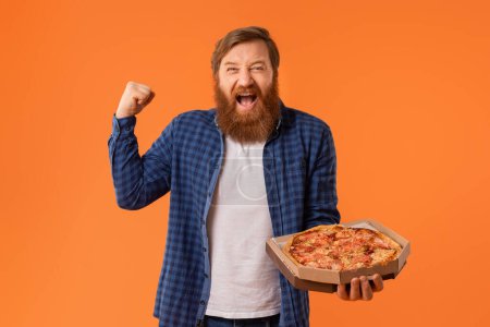 Photo for Joyful man with red hair and beard holds pizza box, shouting and gesturing yes emotionally, standing against orange backdrop in studio. Concept of junk food overeating, pizza delivery - Royalty Free Image