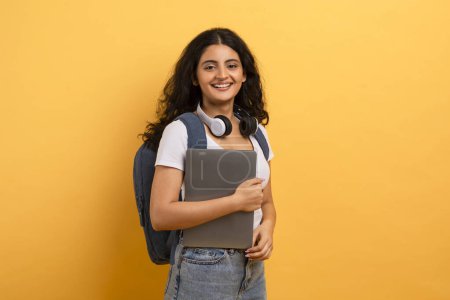 Photo for Happy student with headphones around neck holding a laptop, portrays a mix of education and technology - Royalty Free Image