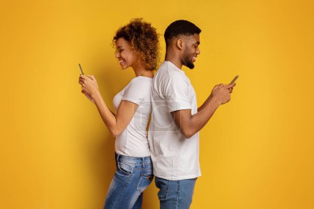 Téléchargez les photos : Un jeune couple afro-américain se tient dos à dos, absorbé dans leurs smartphones, symbolisant les problèmes de communication modernes sur un fond jaune vif - en image libre de droit