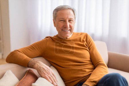 Téléchargez les photos : Confortable gentleman plus âgé se prélassant sur un canapé, souriant largement dans une pièce avec une sensation légère et aérée - en image libre de droit