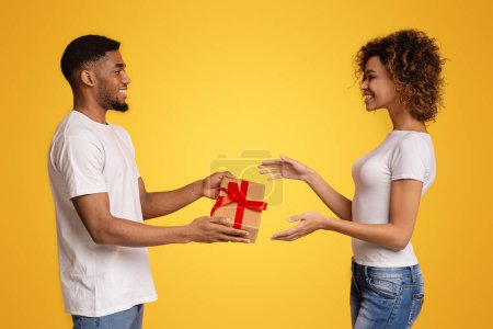 Photo for African American young man presents a red ribbon-wrapped gift box to a delighted young woman against a yellow background - Royalty Free Image