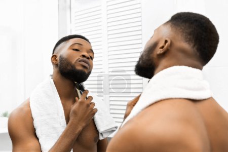 Téléchargez les photos : Gros plan de l'homme afro-américain se toilettant la barbe avec un rasoir électrique, pratique de soins personnels - en image libre de droit