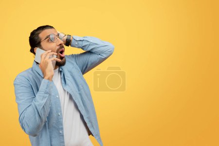 Photo for A perplexed young man in a casual denim shirt talking on phone and touching his head in confusion or forgetfulness against a bright yellow background - Royalty Free Image