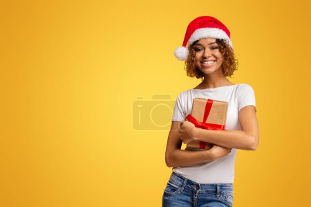 Smiling young African American woman in casual clothes and Santa hat holding a Christmas present, isolated on a yellow background