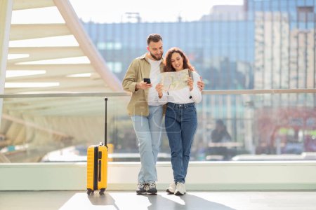 Loving couple holding paper map and using phone, standing at airport with yellow suitcase