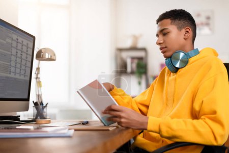 Photo for An attentive young man in a yellow hoodie reads a document sitting at his office desk with headphones - Royalty Free Image