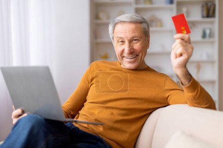 Téléchargez les photos : Un vieil homme souriant brandit une carte de crédit à côté de son ordinateur portable, se livrant probablement à un achat en ligne ou bancaire - en image libre de droit