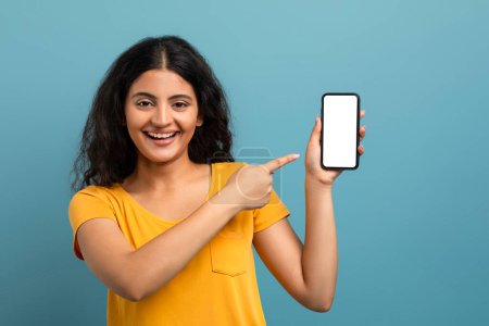 Cheerful woman pointing at a blank smartphone screen with a big smile on a teal background