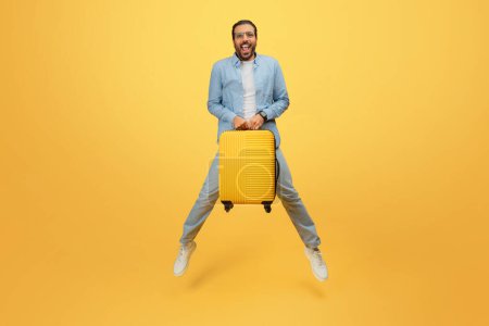 Photo for Happy indian man in denim clothing excitedly grips a yellow rolling suitcase against yellow backdrop - Royalty Free Image