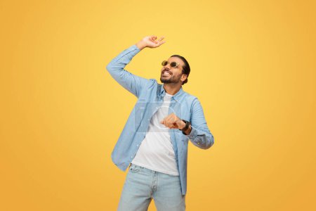 A carefree young indian man with a beard and sunglasses dances joyfully, wearing a light-blue shirt and jeans against a vibrant yellow background