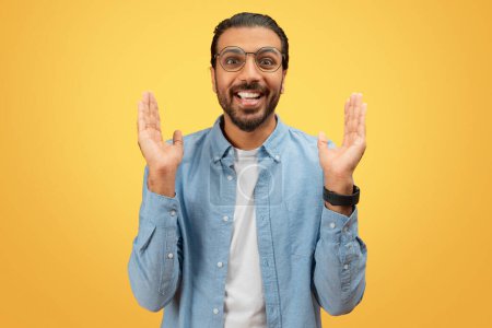 Ein begeisterter indischer Mann mit Brille gestikuliert, um etwas Großes vor einem leuchtend gelben Hintergrund anzudeuten