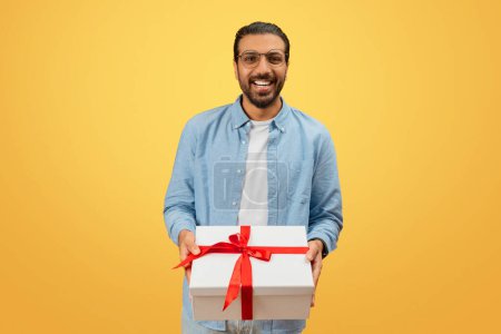 Hombre indio alegre en gafas y una camisa azul que presenta una caja de regalo blanca con una cinta roja