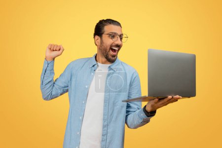 Téléchargez les photos : Homme indien enthousiaste célébrant le succès ou la victoire avec un ordinateur portable ouvert sur fond jaune - en image libre de droit