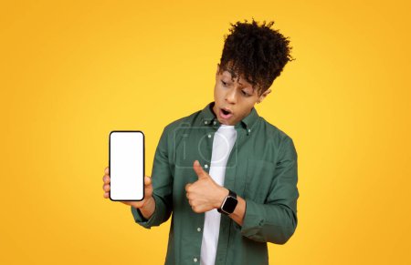 Foto de Joven hombre negro asombrado mostrando una pantalla de teléfono inteligente en blanco, expresando incredulidad, sobre un fondo amarillo vibrante - Imagen libre de derechos