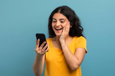 An excited woman in a yellow t-shirt is looking at her phone screen, laughing with a surprised look on a teal background