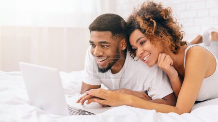 Glückliches afrikanisch-amerikanisches Paar in lässiger Kleidung lächelt beim Betrachten oder Lesen von Inhalten auf einem Laptop im Bett