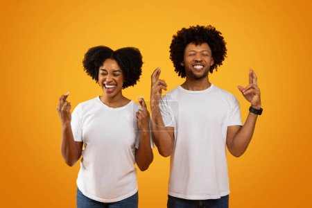 Téléchargez les photos : Superstitieux jeune couple afro-américain croisé les doigts et souriant les yeux fermés isolé sur fond de studio jaune. excité noir homme et femme faire souhait, espoir pour la chance - en image libre de droit