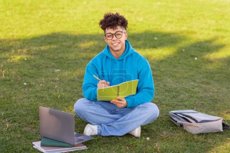 A young brazilian guy wearing eyeglasses writes in a notebook while sitting on grass with laptop and backpack