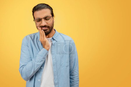 An uncomfortable indian man in glasses holds his cheek, hinting at a toothache or discomfort, against a uniform yellow backdrop