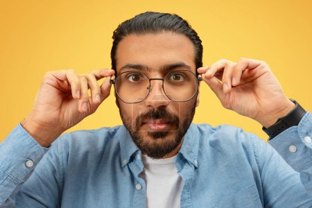Foto de Un hombre indio enfocado ajusta intensamente sus gafas mientras mira hacia adelante sobre un fondo amarillo - Imagen libre de derechos