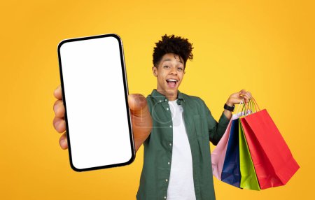 Riendo joven hombre negro sosteniendo un teléfono inteligente con una pantalla de maqueta y bolsas de compras