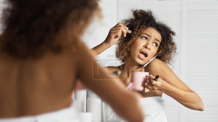 Chica afroamericana con prisa ponerse maquillaje, beber café y hablar por teléfono simultáneamente delante del espejo en el baño. Mañana de concepto de mujer moderna