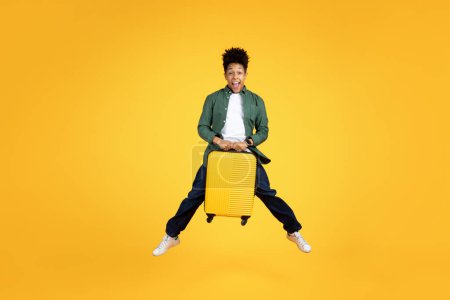 Foto de Un joven afroamericano extático salta con una maleta de color amarillo brillante, expresando alegría y la emoción de viajar sobre un fondo amarillo - Imagen libre de derechos