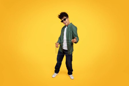 Photo for A young black guy depicted mid-motion against a vivid yellow backdrop, sporting a green shirt and denim jeans - Royalty Free Image