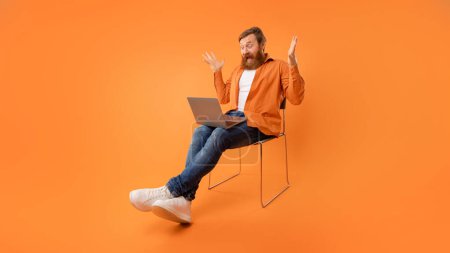Téléchargez les photos : Redhaired homme barbu regarde ordinateur portable et crier avec une expression choquée, éprouver des problèmes pendant la websurf et le travail en ligne, assis avec l'ordinateur dans la chaise au-dessus du mur orange. Panorama - en image libre de droit