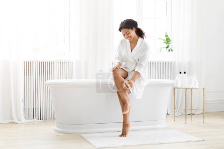 Téléchargez les photos : Une heureuse femme afro-américaine en robe assise sur une baignoire blanche dans une salle de bain lumineuse, moderne et propre, massant ses jambes avec une brosse - en image libre de droit
