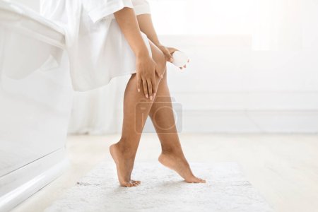 Geschnitten von einer Frau in einem weißen Gewand trägt Feuchtigkeitscreme auf ihre Beine auf und unterstreicht das Konzept der Hautpflege und Selbstpflege zu Hause