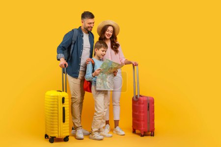 Téléchargez les photos : Une joyeuse famille de trois personnes debout sur un fond jaune, regardant une carte ensemble, avec des valises de couleur vive prêtes pour le voyage - en image libre de droit