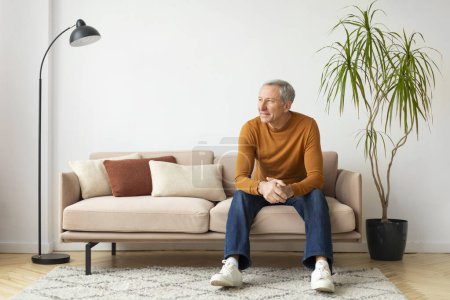 Hombre mayor sentado cómodamente en un sofá en medio de almohadas de tonos cálidos, con una planta en maceta en el fondo
