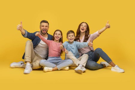 Téléchargez les photos : Une famille joyeuse de quatre personnes avec deux enfants et parents abandonnant les pouces, tous souriants sur un fond jaune vif - en image libre de droit