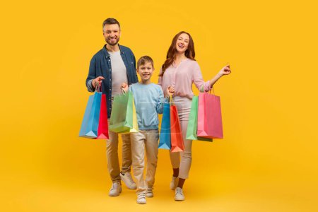 Téléchargez les photos : L'image montre une famille heureuse avec deux adultes et un enfant tenant des sacs à provisions colorés, posant sur un fond jaune vif - en image libre de droit