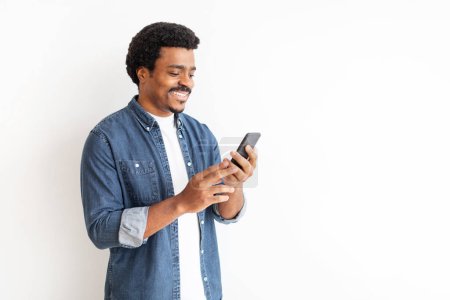 Téléchargez les photos : Joyeux jeune homme noir en denim s'engageant avec smartphone, affichant un sourire chaleureux dans un espace bien éclairé, illustrant la connectivité et la communication moderne, debout sur fond de mur blanc - en image libre de droit