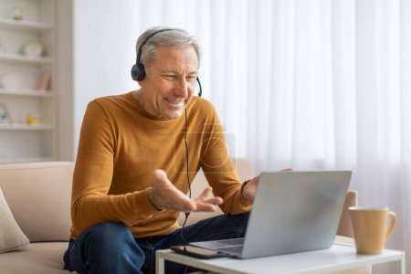 Photo for Senior man wears headphones and actively speaks in a video call in a well-lit home environment, using laptop - Royalty Free Image