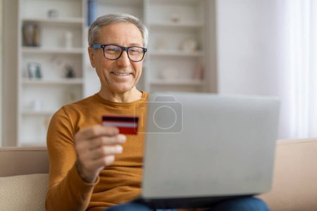 Foto de Anciano sosteniendo una tarjeta de crédito mientras usa una computadora portátil, lo que sugiere compras en línea o banca desde casa - Imagen libre de derechos