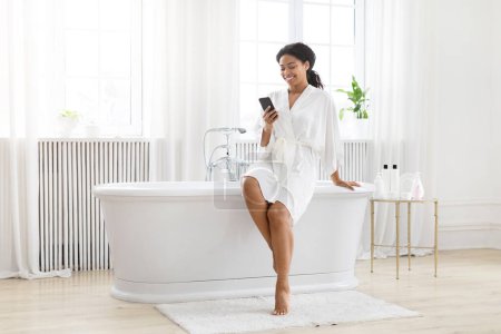 Téléchargez les photos : Femme afro-américaine profite d'un moment de détente dans une salle de bain lumineuse, en utilisant son smartphone près de la baignoire - en image libre de droit