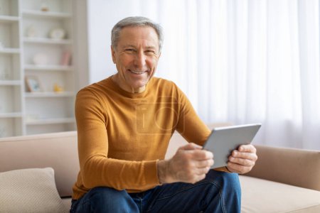 Ein älterer Mann mit einem fröhlichen Lächeln blickt auf einen digitalen Tablet-Bildschirm in einem gemütlichen Zuhause.