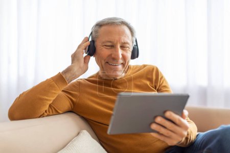 Photo for A happy senior man comfortably uses a tablet while wearing headphones, likely enjoying media at home - Royalty Free Image