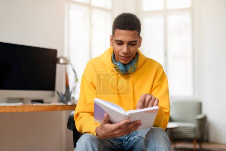 A young man in a yellow hoodie reads from a notebook, seated comfortably at a home office space