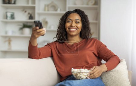 Photo for Cheerful black woman sitting on a couch with popcorn, enjoying television in a cozy living room, african american female relaxing at home - Royalty Free Image