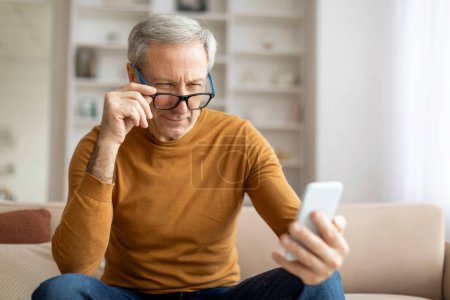 Foto de Anciano caballero toma una pausa contemplativa mientras se conecta con su teléfono inteligente en casa, ajustando gafas - Imagen libre de derechos