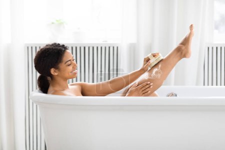 Téléchargez les photos : Une jeune femme noire profite d'un moment de détente dans une baignoire blanche, se brossant la jambe, entourée d'une salle de bain sereine - en image libre de droit