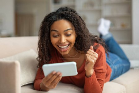 Téléchargez les photos : Joyeuse femme noire allongée sur le canapé réagit à gagner des nouvelles sur son téléphone portable avec une pompe poing gaie, femme afro-américaine célébrant le succès à la maison - en image libre de droit