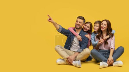 A joyful family of four sitting closely, the father points away while everyone looks cheerful against a yellow background
