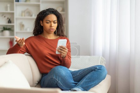 Téléchargez les photos : Une jeune femme noire est assise sur un canapé, regardant perplexe devant son smartphone, une femme afro-américaine souffrant d'escroquerie en ligne, assise sur un canapé à la maison - en image libre de droit