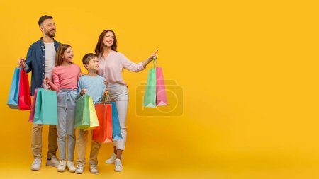 Téléchargez les photos : Parents de famille ludiques et enfants exhibant des sacs à provisions, debout sur jaune, pointant vers l'espace de copie - en image libre de droit