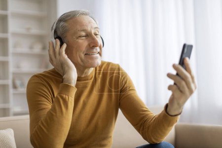 Téléchargez les photos : Heureux senior homme gestes avec la main alors que la vidéo appelant sur un smartphone dans un cadre chaleureux, en utilisant casque - en image libre de droit